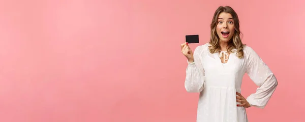 Porträtt av glad glada snygg blond flicka i vit klänning, att vara imponerad och nöjd med cool banktjänst, visar kreditkort, leende kamera, stående rosa bakgrund — Stockfoto