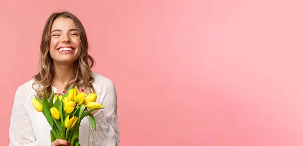 Conceito de primavera, felicidade e celebração. Close-up de menina europeia loira feliz e despreocupada receber belo buquê de flores, segurando tulipas amarelas, sorrindo e rindo, fundo rosa — Fotografia de Stock