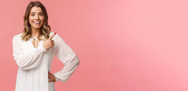 Upbeat chica rubia caucásica bien parecido en vestido blanco, señalando esquina superior derecha y sonriendo a la cámara con interesado, cara curiosa discutir descuentos especiales de primavera promocional, fondo rosa — Foto de Stock