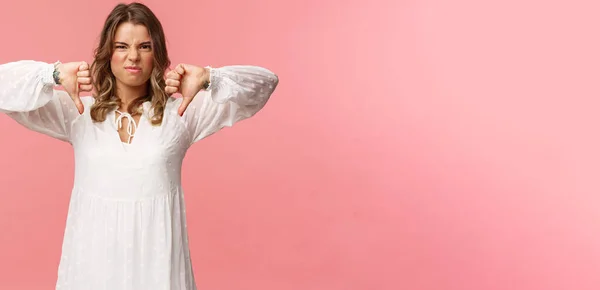 Porträtt av kräsen ung blond kaukasiska kvinna i vit klänning över rosa bakgrund uttrycker ogillar, visa tummen ner och grimma i motvilja, besviken med dålig kvalitet — Stockfoto