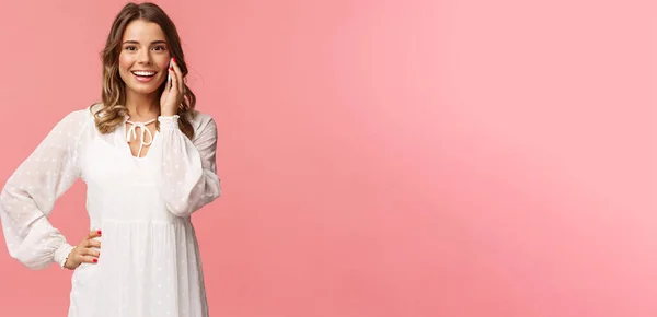 Портрет радостной симпатичной блондинки в белом платье, разговаривающей по телефону, держащей смартфон рядом с ухом и смотрящей оптимистичной камерой, улыбающейся обсуждающей свидания друзей, стоящей на розовом фоне — стоковое фото
