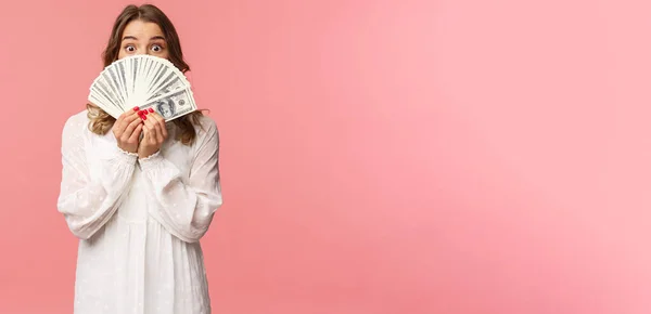 Portret zdumionej i podekscytowanej uroczej blondynki w białej sukience, trzymającej dolary nad twarzą, patrzącej spod gotówki przed kamerą z zaskoczonym wyrazem twarzy, stojącej na różowym tle — Zdjęcie stockowe