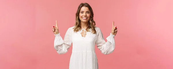 Retrato de confiante bela jovem loira em vestido bonito branco, apontando os dedos para cima na propaganda superior, olhando para a câmera com sorriso radiante, de pé fundo rosa — Fotografia de Stock