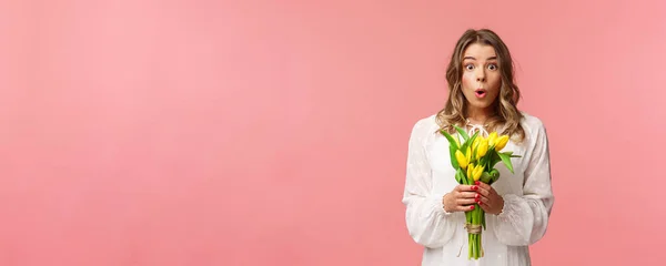Férias, beleza e conceito de primavera. Retrato de menina loira surpreso e surpreso em vestido branco, segurando tulipas amarelas, receber flores sendo divertido e feliz, de pé fundo rosa — Fotografia de Stock