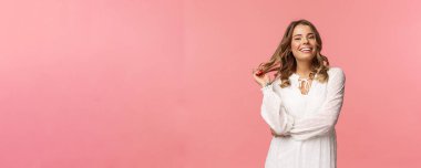 Güzel ve kendine güvenen genç, sarışın, hoş, şık, beyaz elbiseli, saçlarına dokunan ve gülümseyen, romantik randevu için giyinen, kadınlar ve güzellik konsepti, pembe arka plan