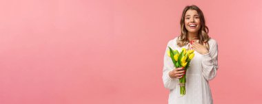 Tatiller, güzellik ve bahar konsepti. Minnettar büyüleyici sarışın kız sarı laleler alıyor, romantik hediye için teşekkür ediyor, iç çekiyor, gülümsüyor, mutlu çiçekler tutuyor, pembe arka plan