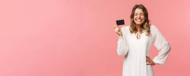 Heyecanlı, tatlı ve aptal, beyaz elbiseli, sarışın bir kızın portresi, kapalı gözlü, kıkırdayan ve gülümseyen mutlu, ilk ödemesini yaptı, kredi kartı tutuyor, bankacılık hizmetini kullanıyor, pembe arka plan