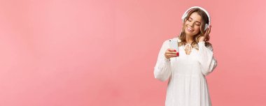 Beyaz elbiseli tatlı ve narin sarışın kız portresi, elinde akıllı telefon, kulaklıkla müzik dinliyor, cep telefonuna gülümsüyor bahar havası için çalma listesi seçiyor, pembe arka plan