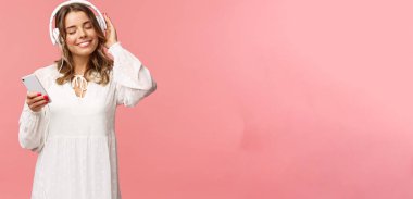 Şefkatli, mutlu ve tatlı sarışın kızın portresi, beyaz elbiseli, kapalı gözlü ve gülümseyen kulaklıkla müzik dinlerken, sıcaklığı ve mutluluğu hissederken, favori parçanın tadını çıkarırken, cep telefonunu tut.