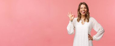 Kısa kıvırcık saçlı mutlu sarışın kadının portresi, işaret dilini göster, tamam de, kabul et ya da onayla, harika ürünü değerlendir, garanti kalite, pembe arka plan