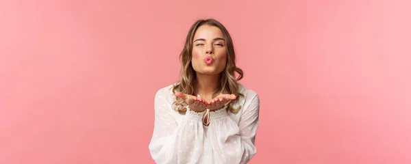 Close-up portret pięknej kobiecej dziewczyny korzystających wiosną piękny dzień, wysyłając pocałunek powietrza przed kamerą, mrugając i składane usta w mwah, trzymać się za ręce w pobliżu ust, różowe tło — Zdjęcie stockowe