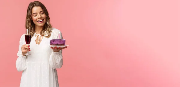 Vacaciones, primavera y concepto de fiesta. Retrato de mujer rubia joven tierna y elegante sosteniendo copa de vino y pastel con vela encendida, sonriendo complacido, celebrando cumpleaños, fondo rosa — Foto de Stock