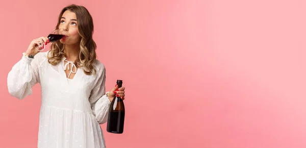 Święta, wiosenna i imprezowa koncepcja. Portret młodej eleganckiej szczęśliwej blondynki, ubranej w białą modną sukienkę, pijącej wino ze szklanki wyglądającej na zadowoloną, degustującej dobry napój, trzymającej butelkę — Zdjęcie stockowe