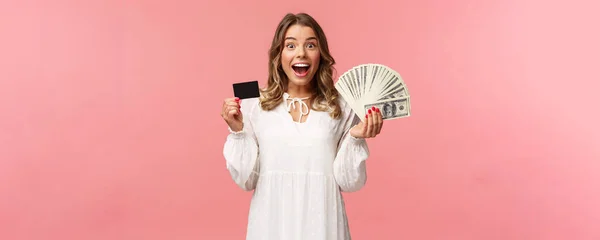 Retrato de feliz menina loira bem-parecido animado em vestido branco, ganhar dinheiro, fez uma boa aposta, fez negócio, segurando dinheiro dólares e cartão de crédito, sorrindo divertido para a câmera, fundo rosa — Fotografia de Stock