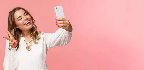 Close-up retrato de alegre linda, menina loira feminina em vestido branco, tomando selfie no telefone celular, fazer kawaii sinal de paz enquanto tirar foto, captura de momento de primavera, fundo rosa — Fotografia de Stock