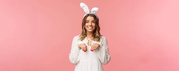假期，春天和派对的概念。可爱娇嫩的金发姑娘模仿兔子的形象，手牵着爪子，戴着兔子耳朵，笑着相机，粉红的背景 — 图库照片