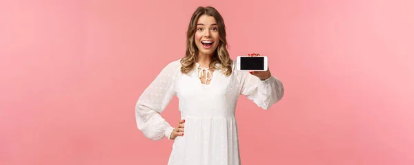 Портрет веселый, оптимистичный привлекательный блондинка в белом платье, показывая дисплей смартфона, держать мобильный телефон горизонтальная улыбающаяся камера поражен, стоять розовый фон — стоковое фото
