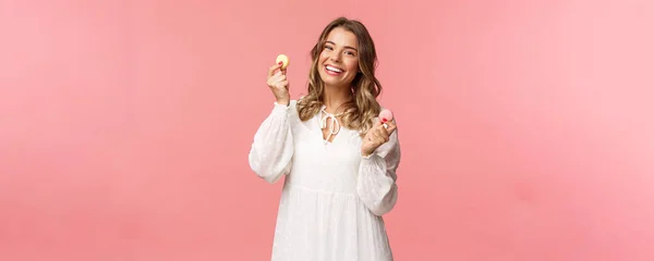 Vacaciones, primavera y concepto de fiesta. Retrato de mujer rubia tierna y encantadora en vestido blanco, bailando alegremente con dos macarrones, sonriendo feliz comiendo delicioso postre, comida sabrosa, fondo rosa — Foto de Stock