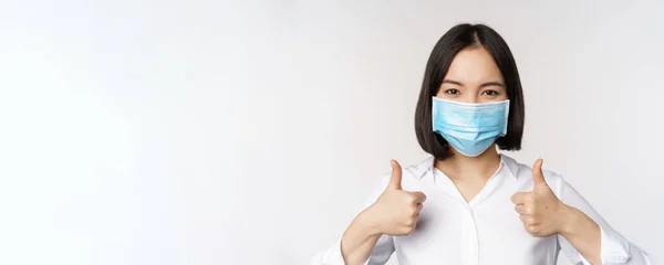 Portrait de dame de bureau asiatique souriante en masque médical, montrant les pouces vers le haut, recommandant smth, debout sur fond blanc — Photo