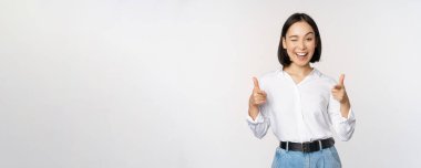Tebrik ederim. Gülümseyen çekici Asyalı kadın, iş kadını mutlu bir yüzle kamerayı işaret ediyor, iltifat ediyor, davet ediyor, beyaz arka planda duruyor.