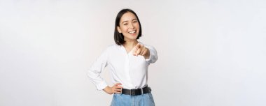 Sensin. Güzel genç Asyalı kadın, şirket müdürü kamerayı işaret ediyor ve gülümsüyor, insanları seçiyor, davet ediyor, beyaz arka planda duruyor.