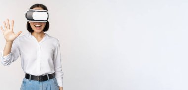 Gülümseyen Asyalı kadın sana el sallıyor, sanal gerçeklik gözlüğü takıyor, kulaklık takıyor, meta evrende sohbet ediyor, beyaz arka planda duruyor.