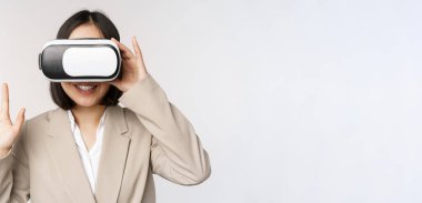 VR chat 'te toplantı. Sanal gerçeklik gözlüklü Asyalı iş kadını, elini kaldırıyor ve merhaba diyor, beyaz arka planda duran birini selamlıyor.