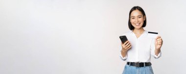 İnternetten alışveriş konsepti. Gülümseyen modern Asyalı kız kredi kartını gösteriyor, cep telefonunu tutuyor, akıllı telefonuyla sipariş veriyor, beyaz arka planda duruyor.