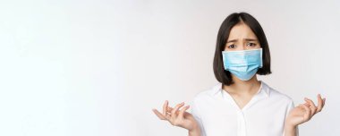 Üzgün ve sefil Asyalı kadın, yüz maskesi takmış, yukarı bakıyor, üzgün bakıyor, beyaz arka planda duruyor.