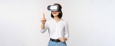 VR kulaklık kullanan Asyalı genç bir kadın. Sanal gerçeklik gözlüklü Koreli kız güler yüzlü, beyaz arkaplanı işaret ediyor.