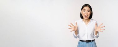 Asyalı kadın kameraya bakar ve panik içinde bağırır. Genç Koreli kız endişeli görünüyor, panikliyor, el sıkışıyor ve bağırıyor, beyaz arka planda duruyor.