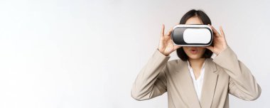 Şaşırmış ofis kadını, Asyalı iş adamı takım elbiseli, kulaklık takıyor, sanal gerçeklik gözlükleriyle smth 'e bakıyor. Etkilenmiş yüz, beyaz arka plan.