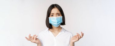 Üzgün ve sefil Asyalı kadın, yüz maskesi takmış, yukarı bakıyor, üzgün bakıyor, beyaz arka planda duruyor.