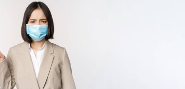 Coronavirus ve iş konsepti. Hüsrana uğramış Asyalı iş kadını, tıbbi yüz maskesi takmış, solu işaret ediyor ve şikayet ediyor, beyaz arka planda duruyor.