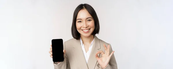 Mujer asiática sonriente mostrando la pantalla del teléfono inteligente y signo de aprobación. Persona corporativa demuestra interfaz de aplicación de teléfono móvil, de pie sobre fondo blanco — Foto de Stock