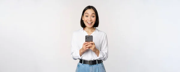 Emocionado mujer asiática sonriendo, reaccionando a la información en el teléfono móvil, sosteniendo el teléfono inteligente y mirando feliz a la cámara, de pie sobre fondo blanco — Foto de Stock