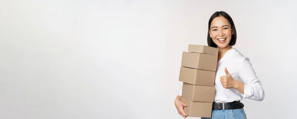 Imagen de la mujer asiática moderna feliz mostrando los pulgares hacia arriba, sosteniendo cajas de entrega mercancías, de pie sobre fondo blanco —  Fotos de Stock