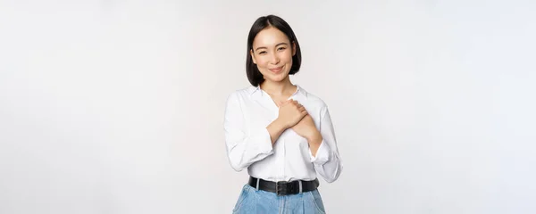 Bild einer fürsorglichen jungen schönen asiatischen Frau, die Hände auf dem Herzen hält, zufrieden und glücklich schaut, in die Kamera lächelt, vor weißem Hintergrund steht — Stockfoto