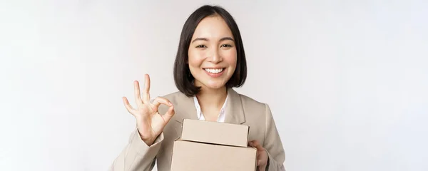 Sorrindo asiático empresária, mostrando bem sinal e caixas com entrega bens, preparar ordem para o cliente, de pé sobre fundo branco — Fotografia de Stock