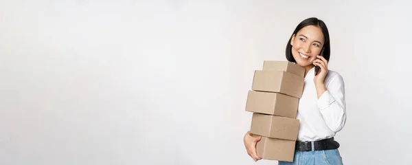 Imagen de joven mujer de negocios asiática contesta llamada telefónica mientras lleva cajas para la entrega, posando sobre fondo blanco —  Fotos de Stock