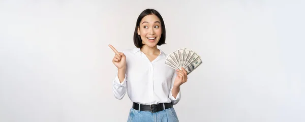 Leende ung modern asiatisk kvinna, pekar på banner annons, hålla kontanter pengar dollar, stående över vit bakgrund — Stockfoto