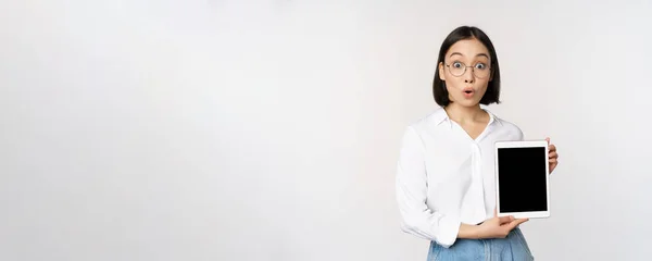 Энтузиастская азиатка, офисный работник в очках, показывающий цифровой экран планшета, демонстрирующий информацию на дисплее гаджета, стоя на белом фоне — стоковое фото