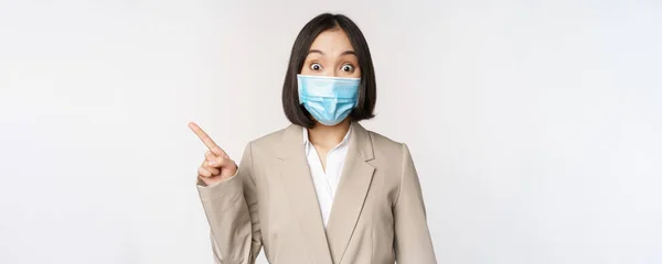 Coronavirus en werkconcept. Portret van vrouw met medisch gezichtsmasker, wijsvinger links, met logo of spandoek, reclame, witte achtergrond — Stockfoto