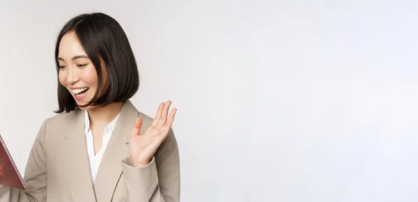 Portrait de sourire asiatique femme d'affaires chat vidéo sur tablette numérique, agitant la main à l'écran du gadget, debout en costume sur fond blanc — Photo