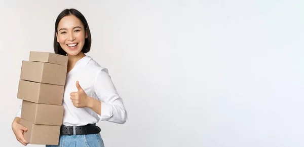 Bild einer glücklichen modernen asiatischen Frau, die Daumen nach oben zeigt, Kisten mit Waren hält und vor weißem Hintergrund steht — Stockfoto