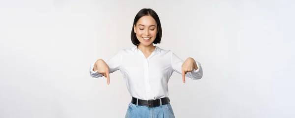 Портрет счастливой азиатки, указывающей пальцем вниз и смотрящей вниз на рекламу, показывающей информационный баннер, рекламу, стоящую на белом фоне — стоковое фото