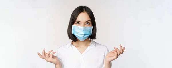 Раздраженная азиатка в медицинской маске для лица, пожимающая плечами и глядя вверх с беспокойным выражением лица, стоящая на белом фоне — стоковое фото