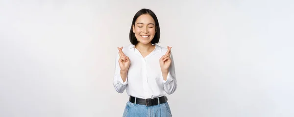 Retrato de mujer joven coreana, chica asiática cruzan los dedos y rezan, pidiendo deseo, anticipando, esperando resultados, de pie sobre fondo blanco — Foto de Stock
