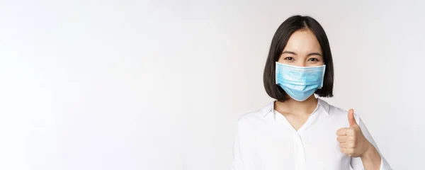 Covid e conceito de saúde. retrato de ásia mulher vestindo médico rosto máscara e mostrando polegares para cima de pé sobre branco fundo — Fotografia de Stock