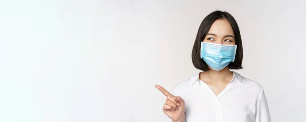 Retrato de bonito ásia mulher no médico rosto máscara, coronavírus proteção, apontando dedo esquerdo e olhando intrigado no vazio cópia espaço, branco fundo — Fotografia de Stock